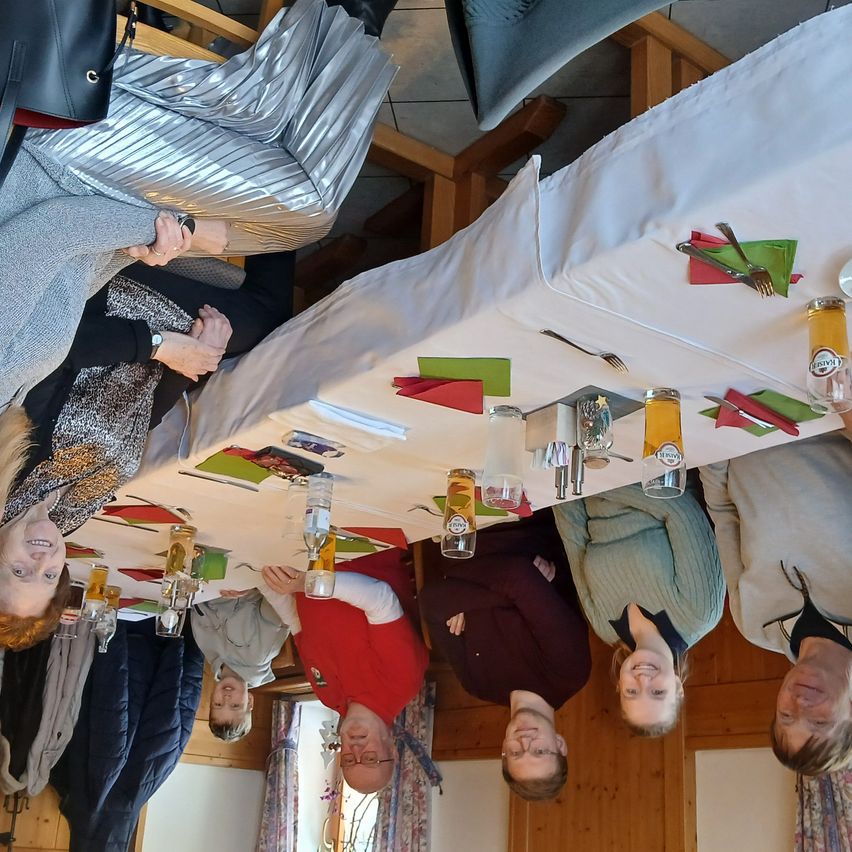 Eine Gruppe von Menschen versammelt sich um einen langen Tisch mit Flaschen und Gläsern in einer festlichen Atmosphäre. Sie lächeln und scheinen für ein Foto zu posieren. Der Tisch ist mit einem weißen Tuch bedeckt und mit roten und grünen Servietten dekoriert.