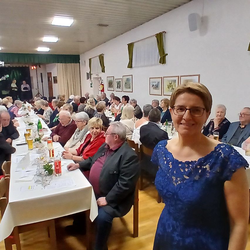 Eine Frau steht in einem Bankettsaal mit vollen Tischen voller älterer Menschen und lächelt in die Kamera. Der Saal hat weiße Wände, grüne Vorhänge und mehrere Bilderrahmen.