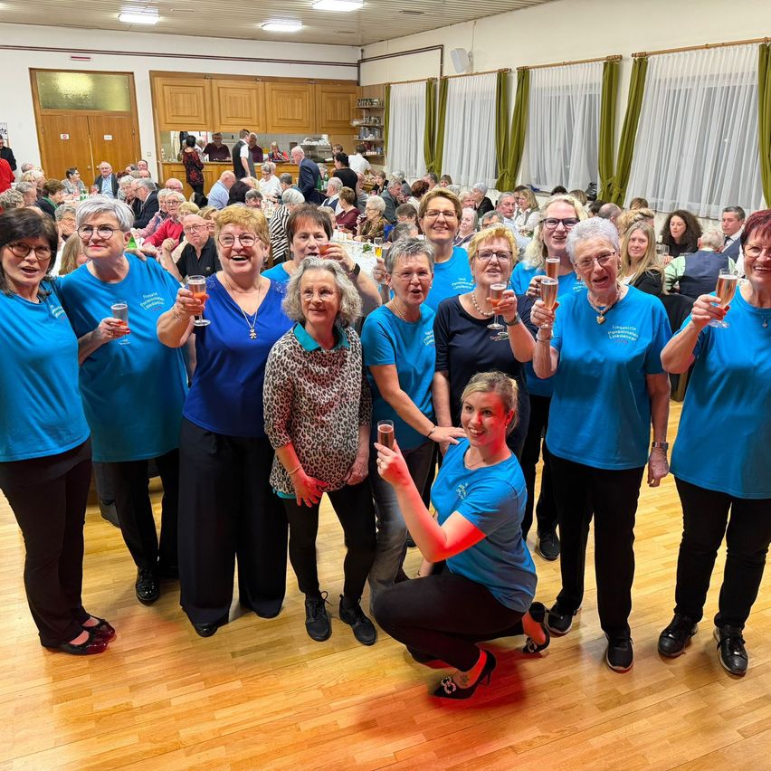 Eine Gruppe von Menschen in blauen Shirts und schwarzen Hosen feiert in einem Raum mit Holzboden, einige halten Getränke. Sie scheinen zu lächeln und posieren für ein Foto.