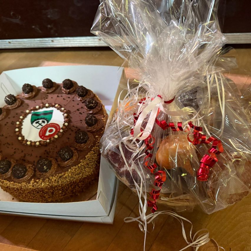 Ein Schokoladenkuchen mit einem Logo in der Mitte befindet sich in einer Schachtel mit einer Plastikabdeckung. Daneben ist eine durchsichtige Plastiktüte mit einem roten Band, die eine Auswahl an Artikeln enthält.