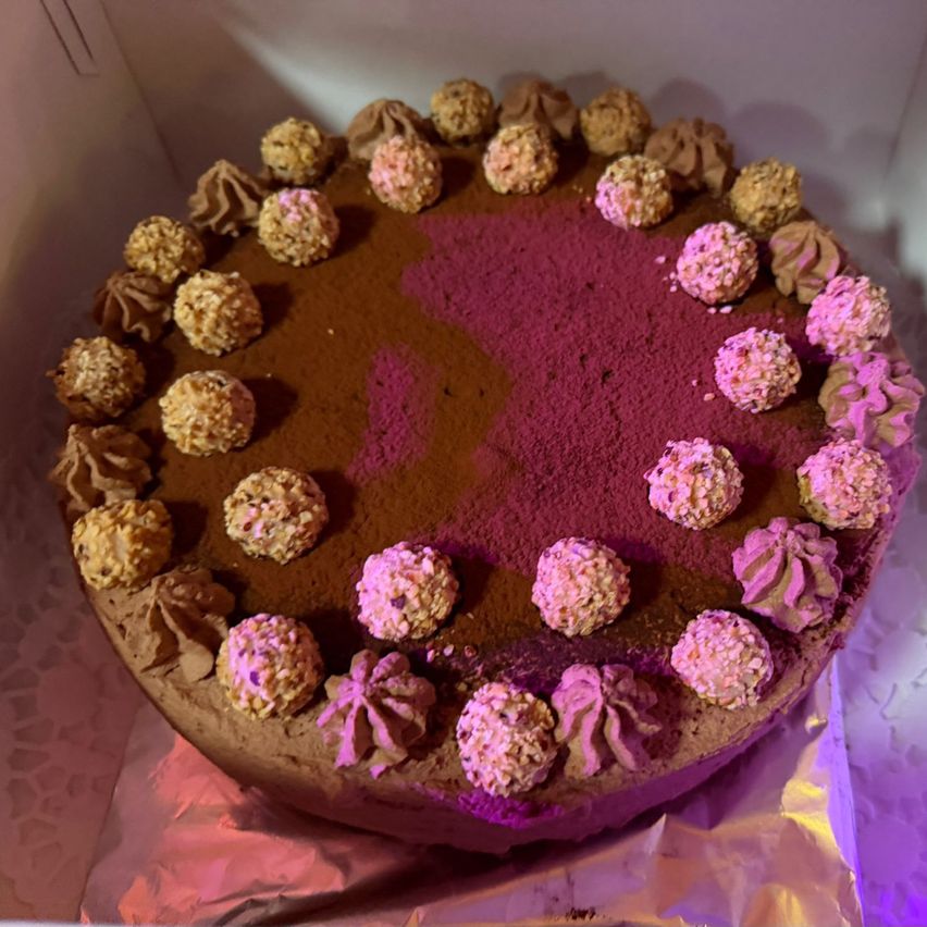 Ein runder Schokoladenkuchen mit lila Mitte, bedeckt mit Schokoladenglasur und gekrönt von kleinen rosa und braunen Kugeln.