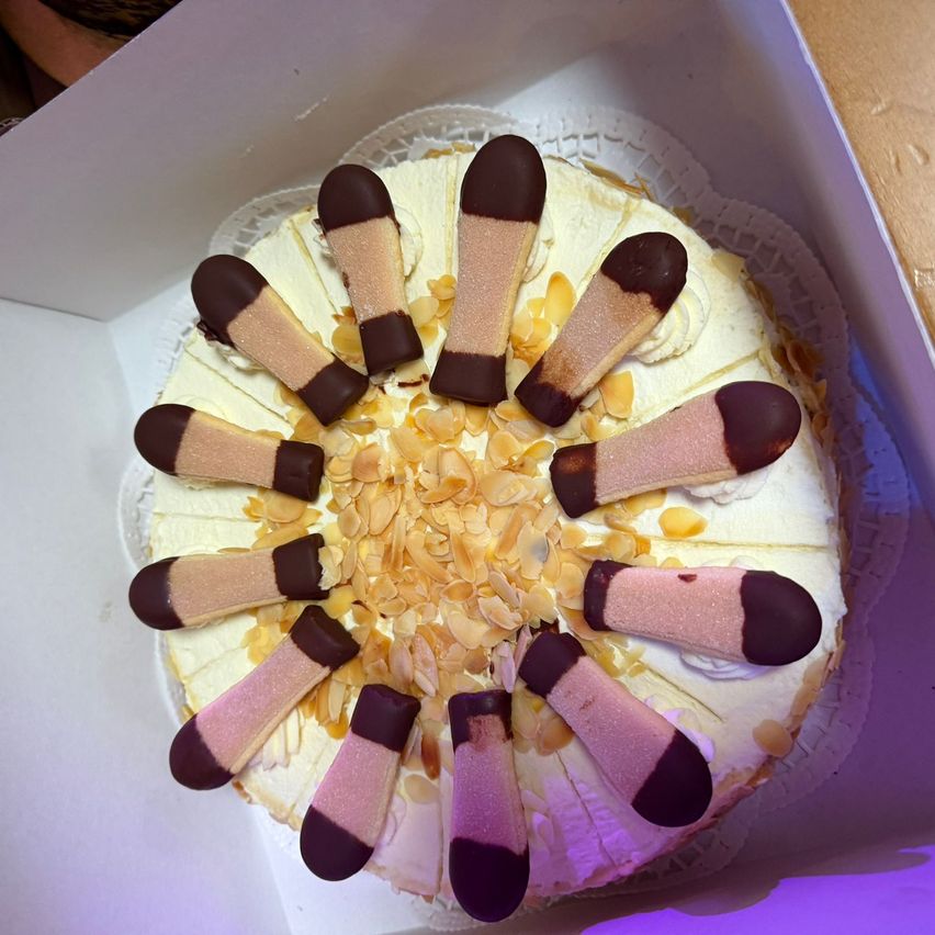 Ein Kuchen mit weißer Basis und rosa und brauner Glasur, verziert mit Mandelscheiben, wird in einer weißen Schachtel präsentiert.