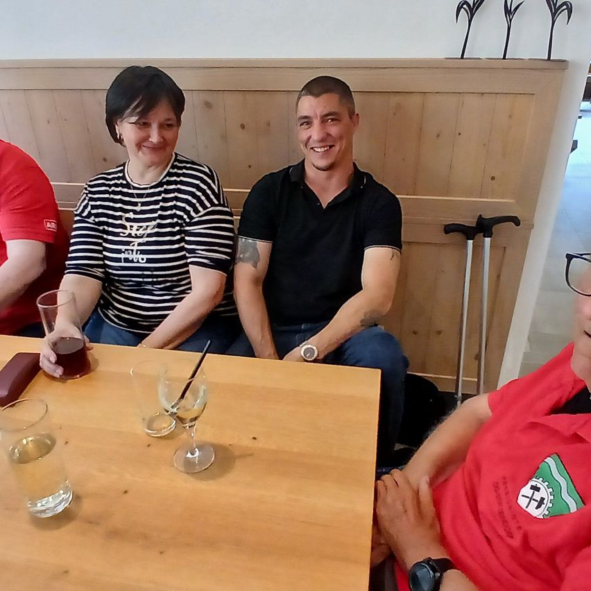 Drei Menschen sitzen in einer Restaurantkabine und posieren lächelnd für ein Foto. Eine Frau in einem gestreiften Shirt und ein Mann in einem schwarzen Shirt. Vor ihnen steht ein Holztisch mit Gläsern und Getränken. Im Hintergrund ist ein Stock zu sehen.