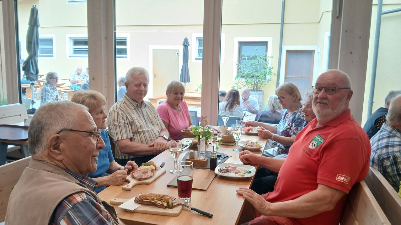 Eine Gruppe von Menschen sitzt in einem Restaurant an einem Tisch und genießt lächelnd ihre Mahlzeiten. Der Tisch ist mit Tellern, Gläsern und verschiedenen Speisen gedeckt. Hinter ihnen ist durch das Fenster ein Gebäude zu sehen.