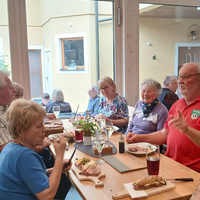 Eine Gruppe älterer Erwachsener sitzt an einem Tisch und genießt eine Mahlzeit. Es gibt Sandwiches, Weingläser und Gewürze auf dem Tisch. Sie scheinen miteinander zu plaudern.