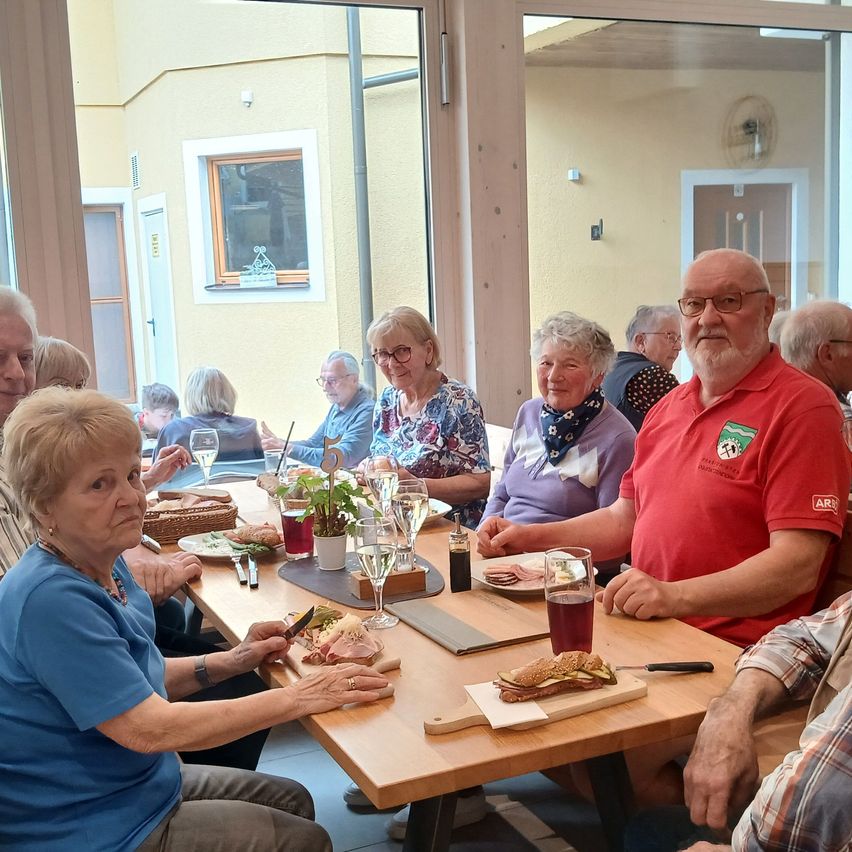 Eine Gruppe älterer Menschen sitzt an einem Tisch und isst und trinkt. Einige tragen Brillen, einer trägt ein rotes Shirt mit Logo. Weingläser und Teller stehen auf dem Tisch. Der Raum hat eine Glastür und Fenster.