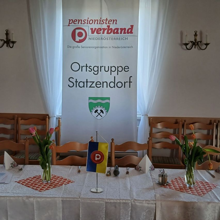 Ein Tisch für eine Veranstaltung, mit zwei Blumenarrangements, einer Flagge und einem Banner mit der Aufschrift 'Pensionistenverband Niederosterreich'. Stühle sind um den Tisch herum angeordnet.