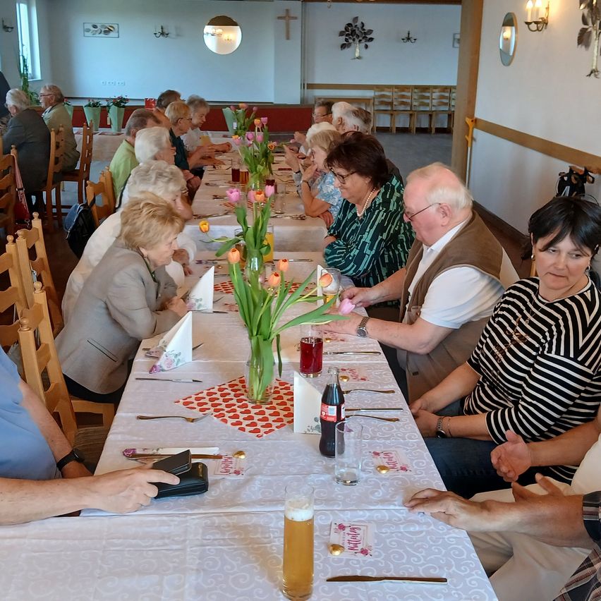 Eine Gruppe älterer Menschen sitzt um einen langen Esstisch und genießt ein Essen. Es gibt Blumen und Getränke auf dem Tisch.