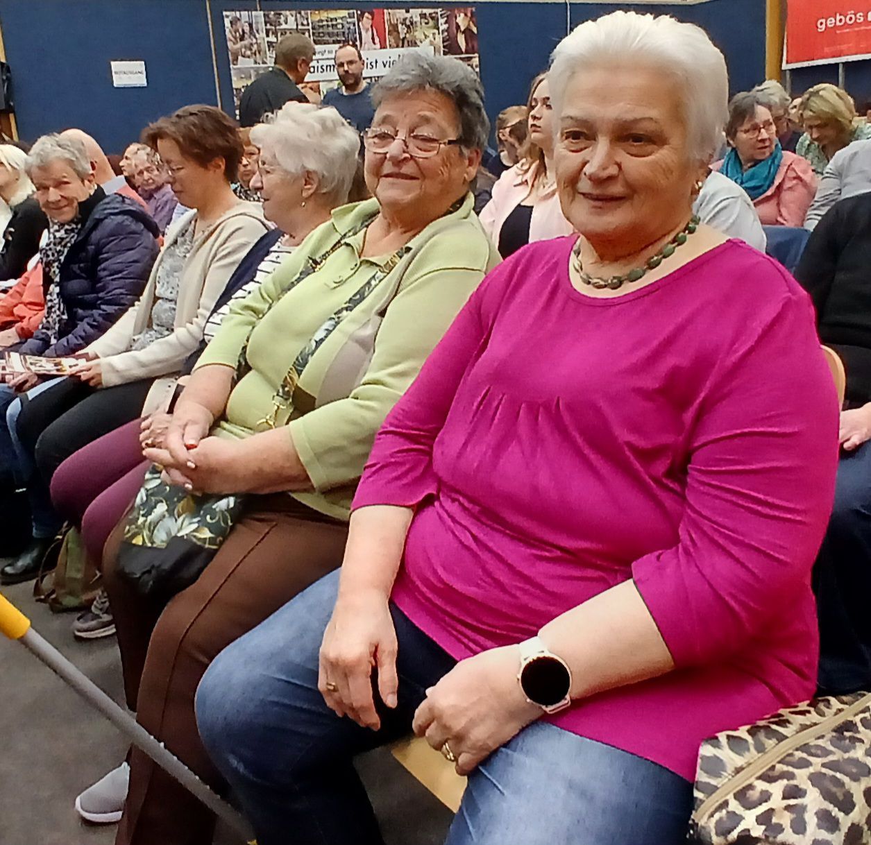 Eine Gruppe älterer Frauen in einem Raum, mit Brillen und verschiedenen Kleidungsstücken. Eine Frau in Pink lächelt, eine andere in Grün hält eine Handtasche.