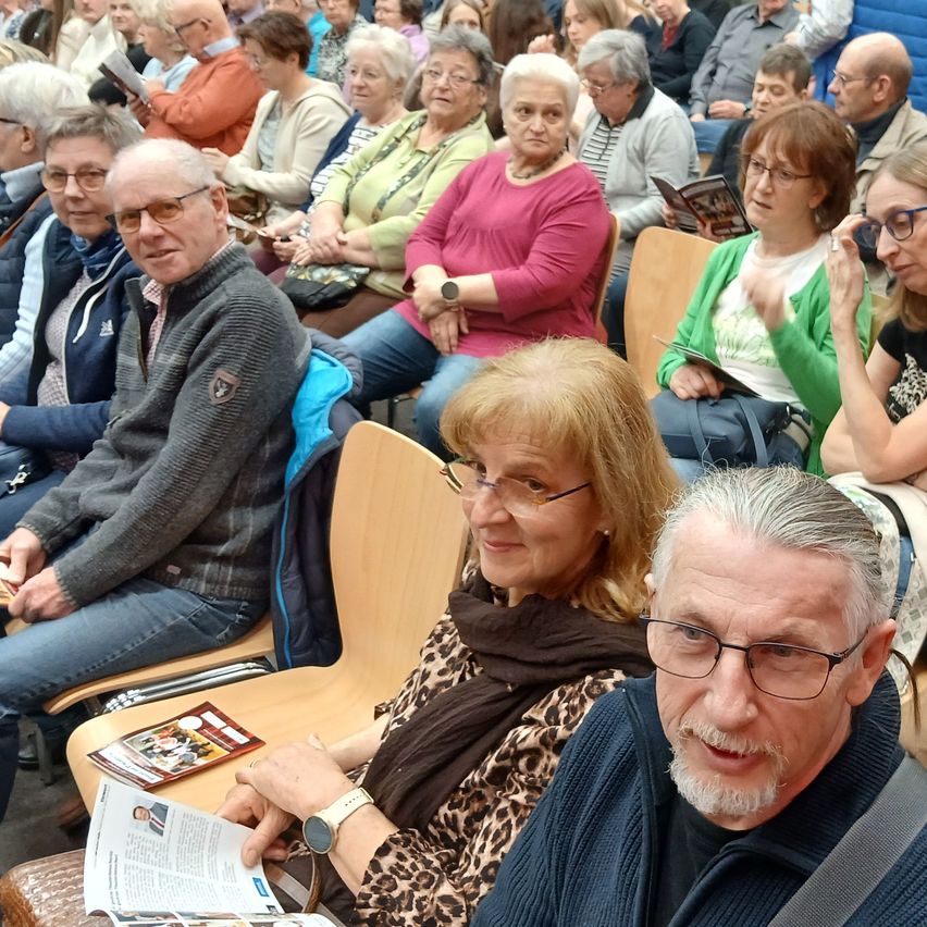Ein Publikum sitzt bei einer Veranstaltung in Stühlen, einige lesen Bücher, andere schauen hinein. Die Frau im Leopardenmuster lächelt in die Kamera.