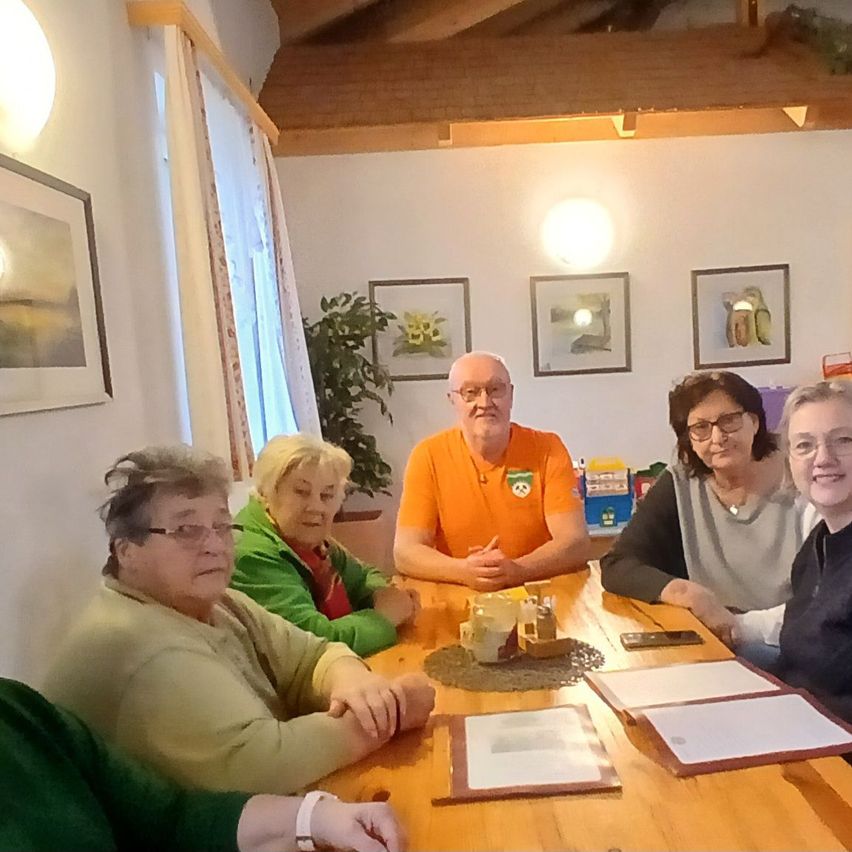 Sechs Menschen sitzen um einen Holztisch mit Papieren und einem Handy. Ein Mann in einem orangefarbenen Shirt und eine Frau in einer grünen Jacke. Sie scheinen eine Besprechung zu haben.