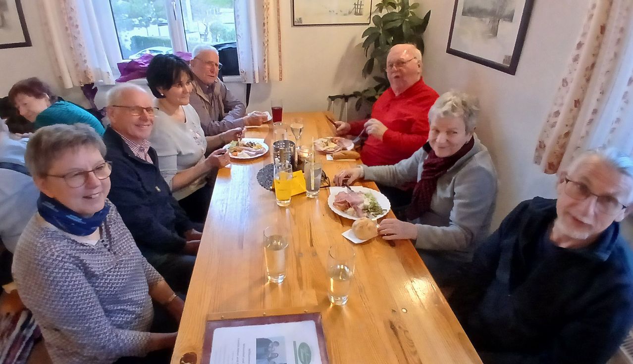 Sechs ältere Menschen sitzen um einen Tisch herum, lächeln und essen. Jeder hat einen Teller mit Essen und Gläser Wasser stehen auf dem Tisch.