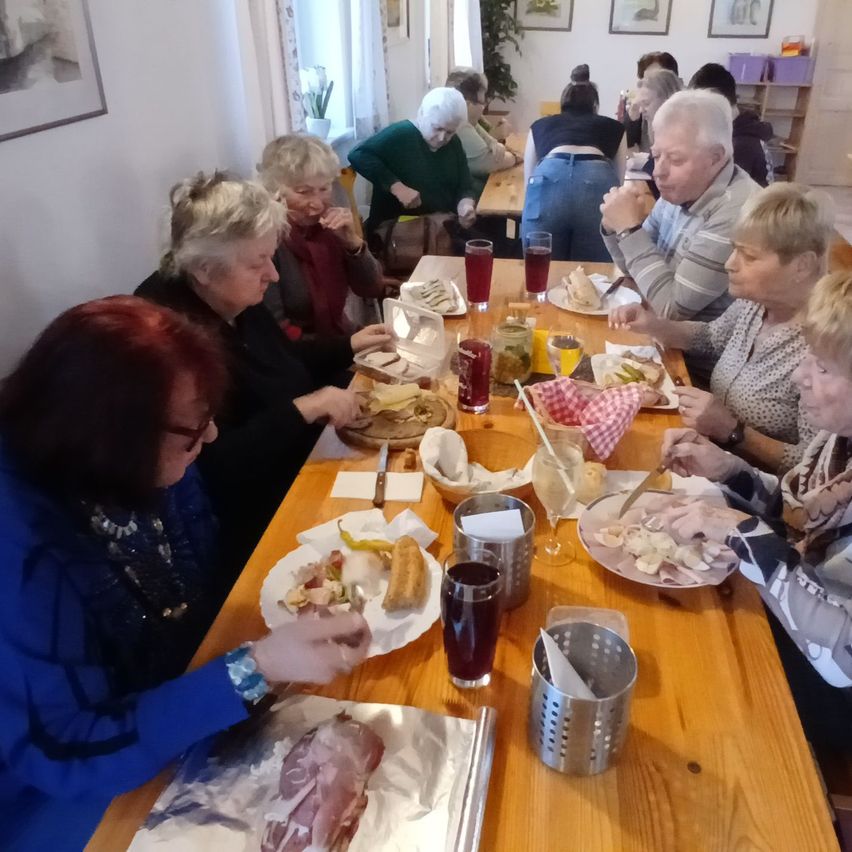 Eine Gruppe älterer Menschen sitzt an einem langen Tisch und genießt eine Mahlzeit. Verschiedene Speisen sind auf dem Tisch, einschließlich Brot, Gemüse und Fleisch. Gläser mit Getränken und Besteck sind ebenfalls vorhanden.