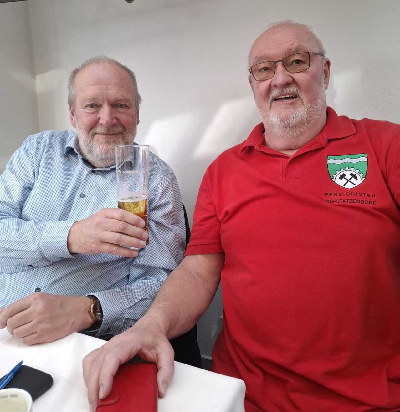 Zwei Männer sitzen an einem Tisch, einer in einem blauen Hemd mit einem Glas Bier in der Hand, der andere in einem roten Hemd mit einem Logo darauf.