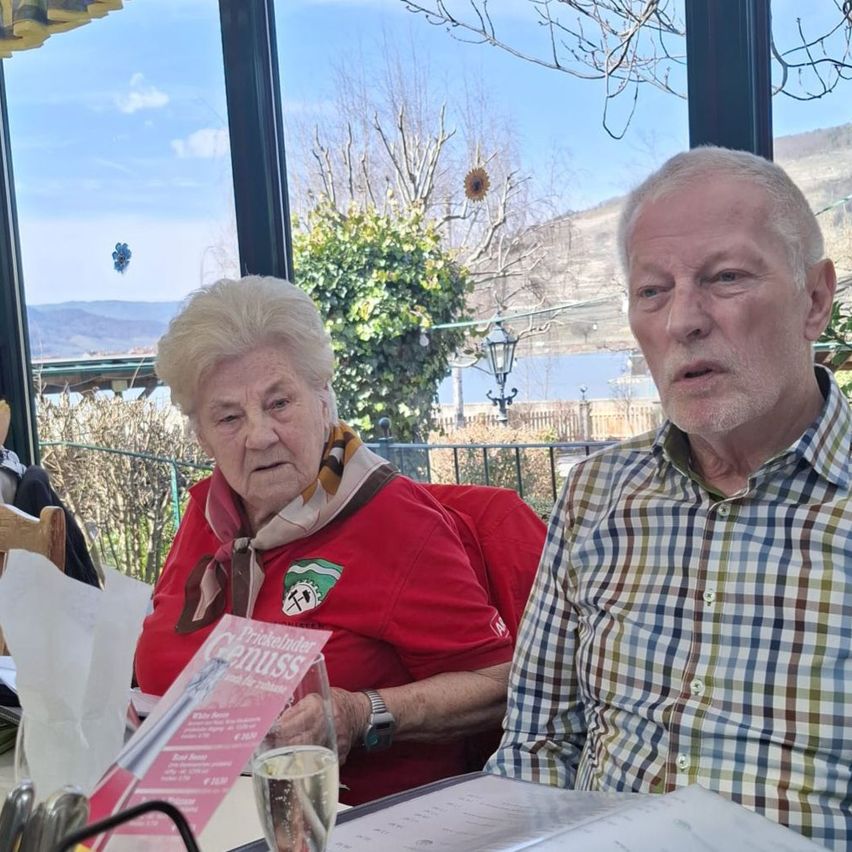 Zwei ältere Personen sitzen an einem Tisch und sehen sich an, in einem Restaurant mit Blick auf Berge und einen See durch die Fenster.