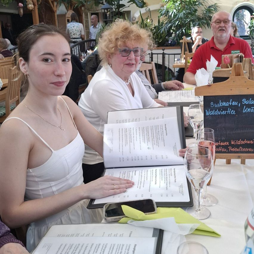 Zwei Frauen und ein Mann sitzen an einem Tisch in einem Restaurant. Die Frau liest eine Speisekarte, während eine andere Frau eine Speisekarte hält. Der Mann sitzt in einem roten Hemd. Es gibt Weingläser und ein Handy auf dem Tisch.