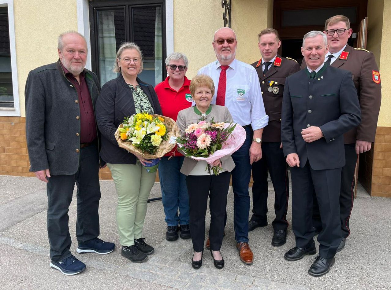 Eine Gruppe Erwachsener steht vor einem Gebäude und hält Blumen. Eine Frau hält zwei Sträuße und andere lächeln für die Kamera.