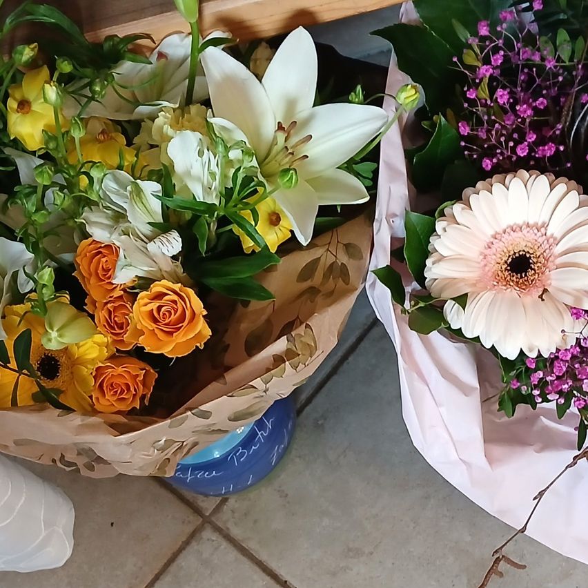 Zwei Blumensträuße liegen auf einem gefliesten Boden. Ein Strauß hat weiße Lilien, orange Rosen und gelbe Blumen, während der andere rosa Blumen und eine weiße Margerite hat.