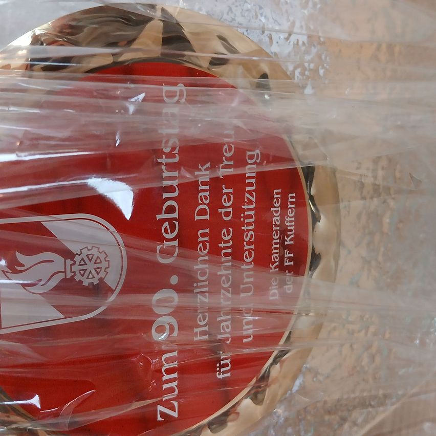 Ein roter runder Plaketten mit goldenem Rand ist zu sehen. Es steht Zum 90. Geburtstag in weiß geschrieben. Auf der linken Seite befindet sich ein Logo und in der Mitte eine Flamme. Die Plakette ist mit durchsichtigem Plastik bedeckt.