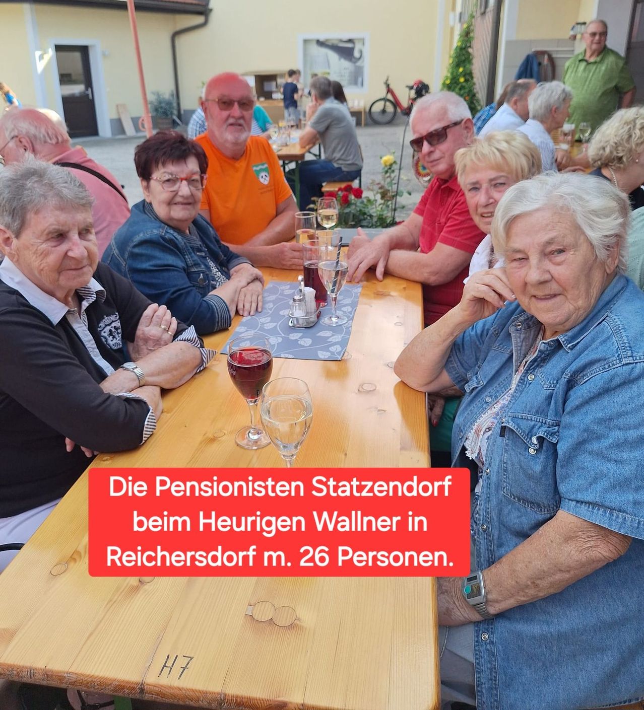 Eine Gruppe älterer Menschen, darunter Männer und Frauen, sitzen an einem Tisch vor einem Restaurant, lächeln und posieren für ein Foto. Der Tisch ist mit Weingläsern, einer Flasche und einem Tuch gedeckt. Das Bild hat einen roten Rand mit weißem Text.