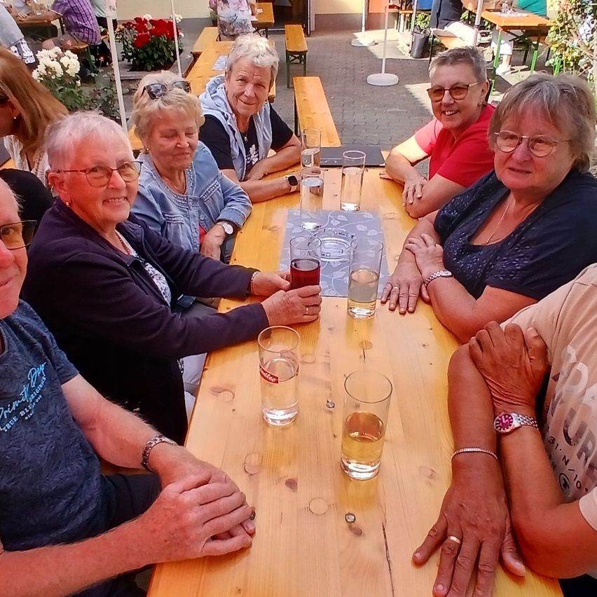 Eine Gruppe älterer Frauen und Männer sitzt an einem Tisch, lächelt und posiert für ein Foto. Sie tragen Brillen und einige haben Halsketten. Der Tisch hat Gläser mit Wasser und eine Speisekarte. Hinter ihnen sind Tische mit sitzenden Menschen und Blumen.