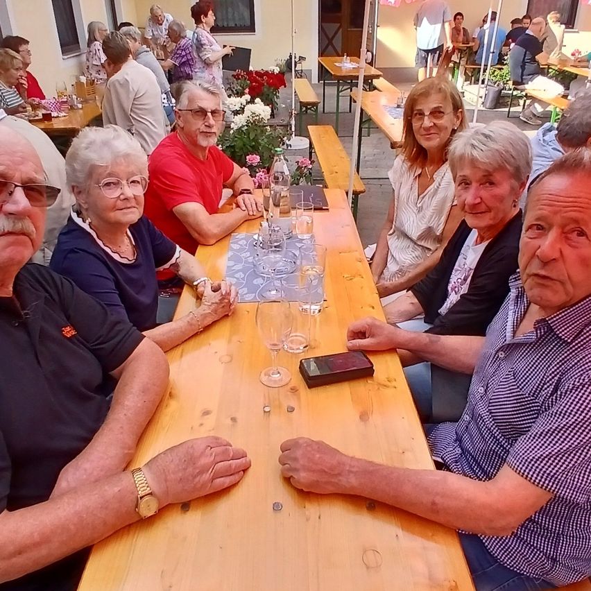 Eine Gruppe älterer Menschen sitzt um einen Tisch in einem Außenrestaurant, einige mit Gläsern und einem Handy auf dem Tisch.
