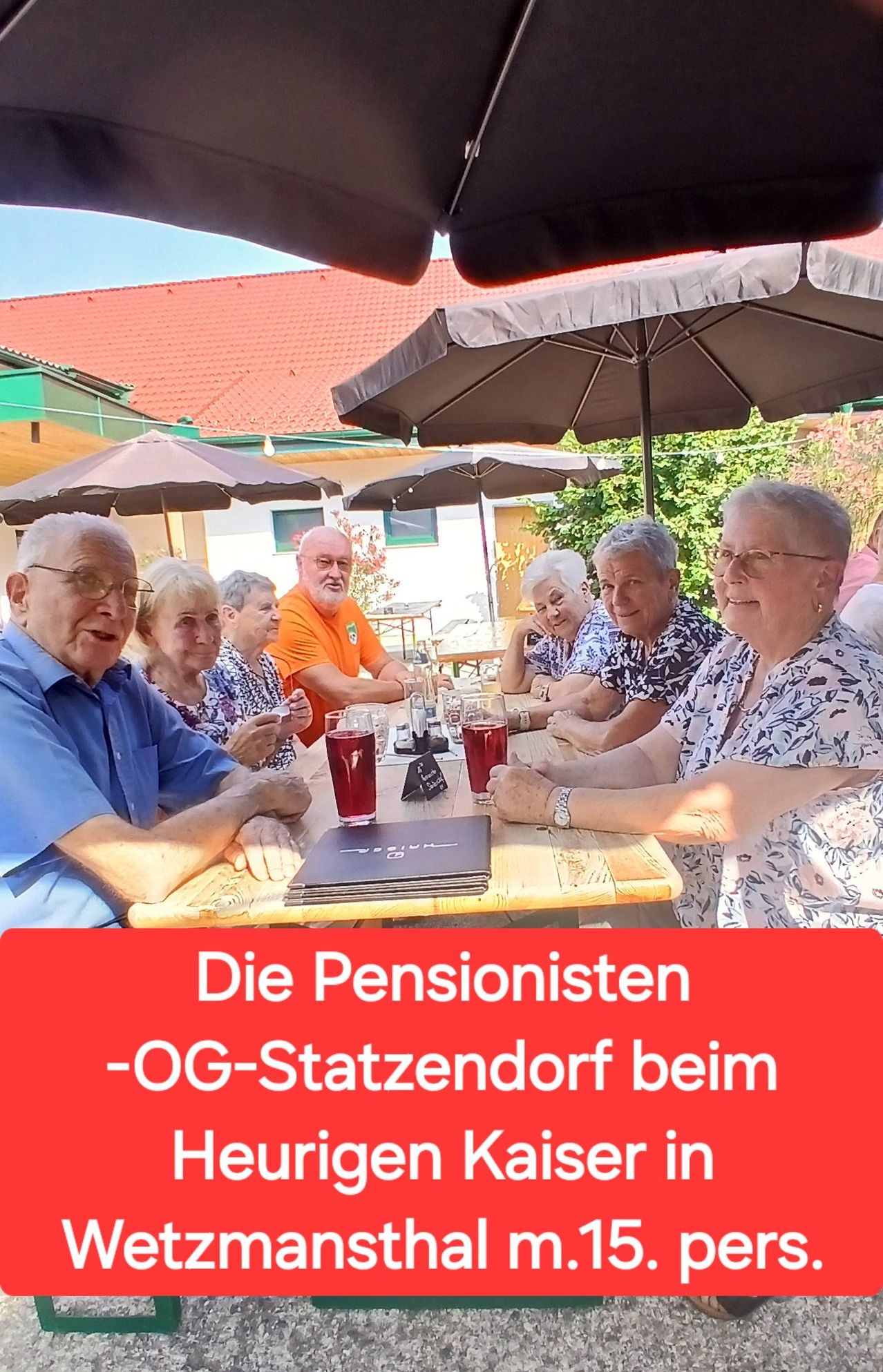 Eine Gruppe älterer Menschen sitzt draußen an einem Tisch unter Schirmen. Sie lächeln und scheinen eine Mahlzeit oder Getränke zu genießen. Der Tisch hat Gläser und eine Speisekarte.