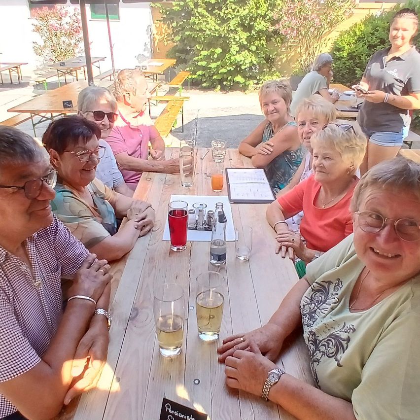 Eine Gruppe älterer Frauen und Männer sitzt an einem Tisch in einem Restaurant im Freien, lächelt und genießt ihre Getränke.