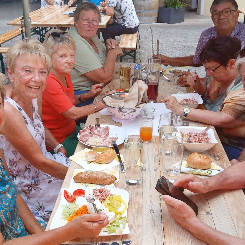 Eine Gruppe älterer Menschen sitzt um einen Tisch in einem Outdoor-Essbereich und genießt ihre Mahlzeiten. Der Tisch ist mit verschiedenen Gerichten, Getränken und Utensilien gefüllt. Die Atmosphäre ist ungezwungen und freundlich.
