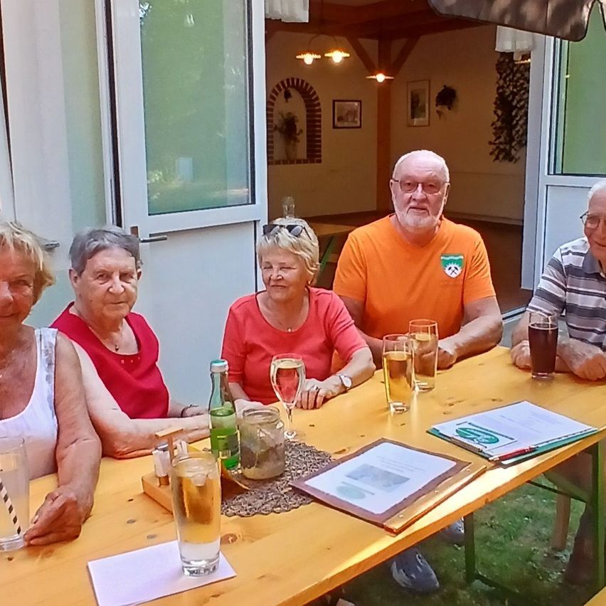 Fünf ältere Erwachsene sitzen draußen an einem Tisch und lächeln für ein Foto. Sie haben Getränke und Snacks vor sich, und eine Person trägt ein leuchtend orangefarbenes Shirt.