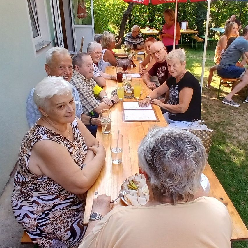 Eine Gruppe älterer Menschen sitzt um einen langen Tisch in einer Außenumgebung, wahrscheinlich bei einer Gartenparty. Sie lächeln und scheinen ihre Mahlzeit und Getränke zu genießen.