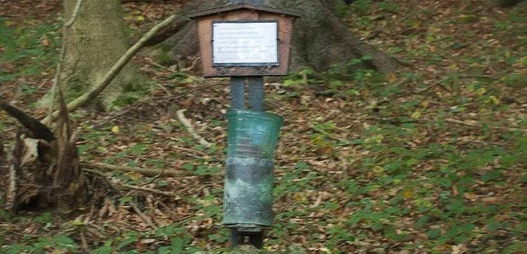 Ein hölzernes Kruzifix ist an einem Baumstamm in einem Wald montiert, darunter ein Metallbehälter. Das Gebiet ist mit abgefallenen Blättern bedeckt.