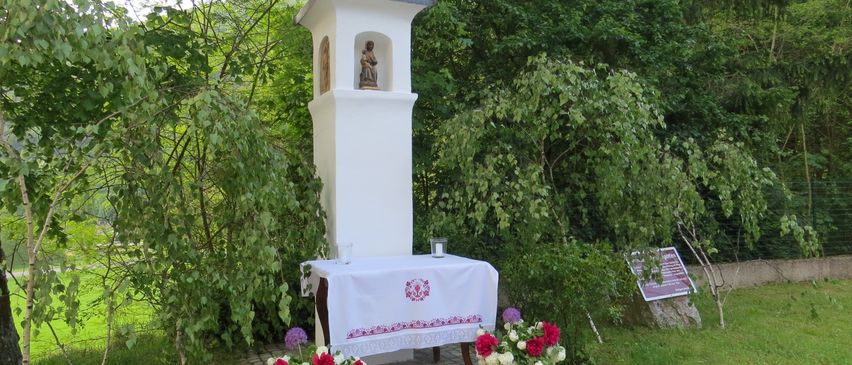 Eine kleine weiße Kapelle mit einem Kreuz auf der Spitze. Eine Statue eines Heiligen ist drinnen. Ein Tisch mit einem Tuch und Blumen steht davor.