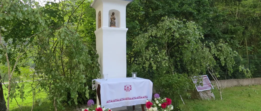 Eine kleine weiße Kapelle mit einem Kreuz auf der Spitze. Eine Statue eines Heiligen ist drinnen. Ein Tisch mit einem Tuch und Blumen steht davor.