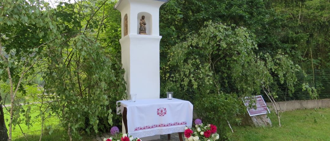 Eine kleine weiße Kapelle mit einem Kreuz auf der Spitze. Eine Statue eines Heiligen ist drinnen. Ein Tisch mit einem Tuch und Blumen steht davor.