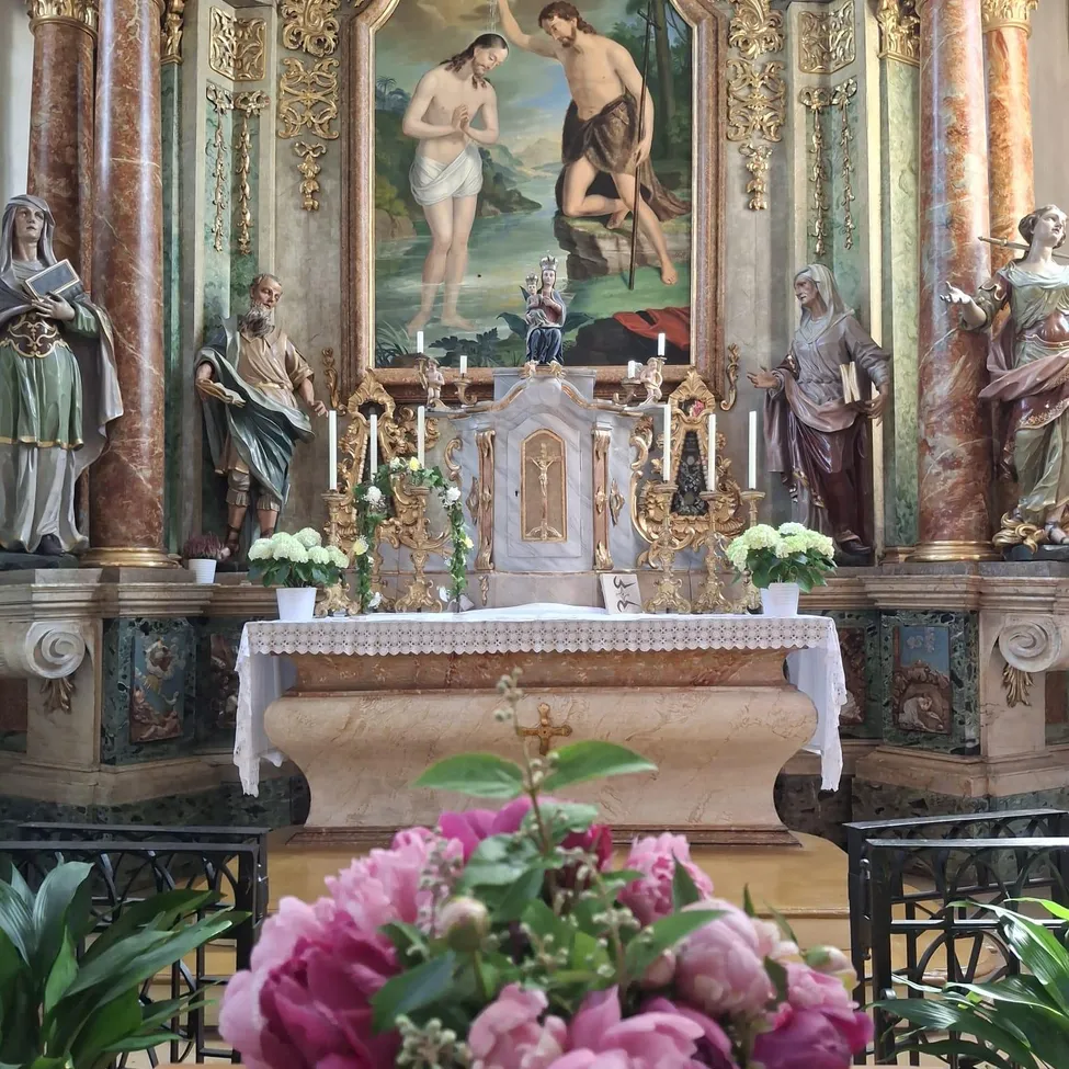 Ein Kirchenaltar mit Statuen und einem Gemälde verziert. Blumenarrangements und Kerzen sind vorhanden.