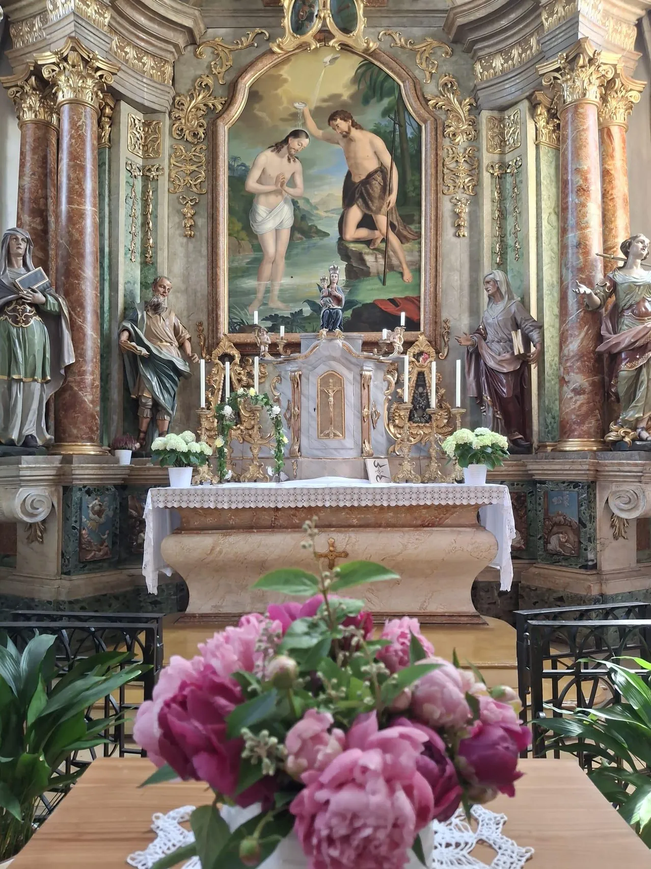 Ein Kirchenaltar mit Statuen und einem Gemälde verziert. Blumenarrangements und Kerzen sind vorhanden.