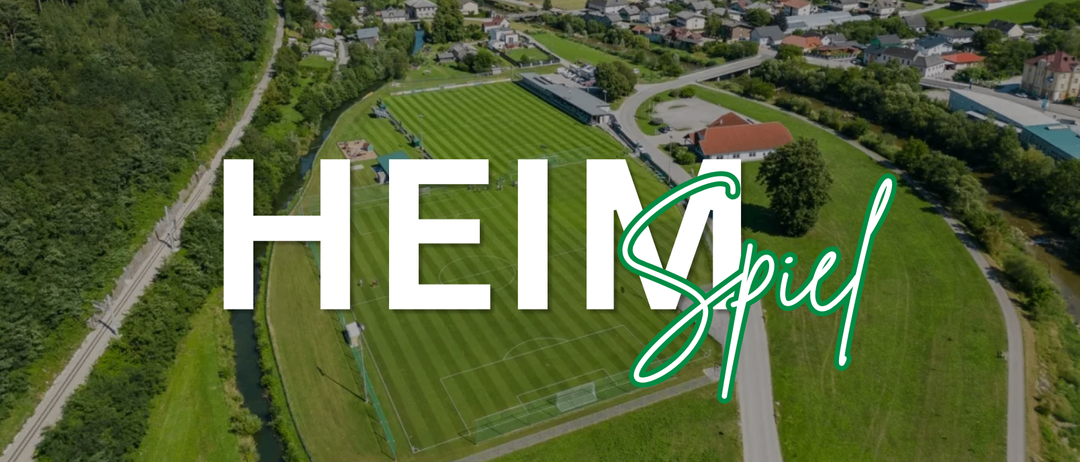 Eine Luftaufnahme eines Fußballfelds mit der Aufschrift 'HEIM', einem Fluss und Häusern in der Ferne.