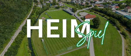 Luftaufnahme eines Fußballfelds mit der Aufschrift 'HEI M' und einem grünen Swoosh. Das Feld ist von Häusern und Bäumen umgeben.