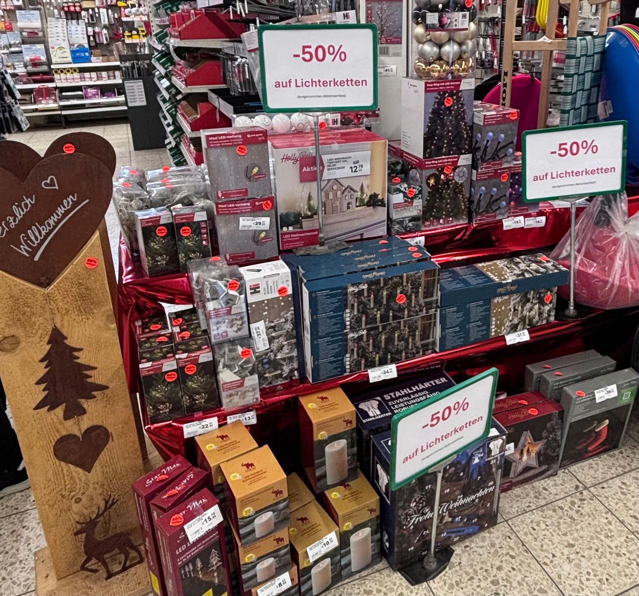 Ein Ladengang zeigt verschiedene Weihnachtslichter, mit einem -50% Rabattschild. Die Regale sind gefüllt mit Schachteln von Lichtern, einige mit 'Holly' und '12' beschriftet, und einem festlichen Holzschild mit Herz und Weihnachtsbaum.