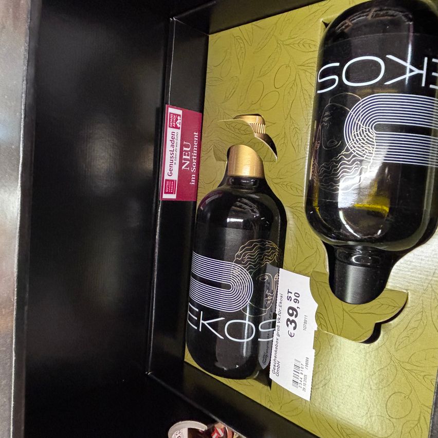 Zwei schwarze Flaschen mit goldenen Verschlüssen in einer schwarzen Box mit goldfarbenem Hintergrund. Ein rotes Etikett auf der linken Flasche sagt 'Neu im Sortiment'. Die rechte Flasche hat ein weißes Etikett mit '39,90' und '10/2021'.