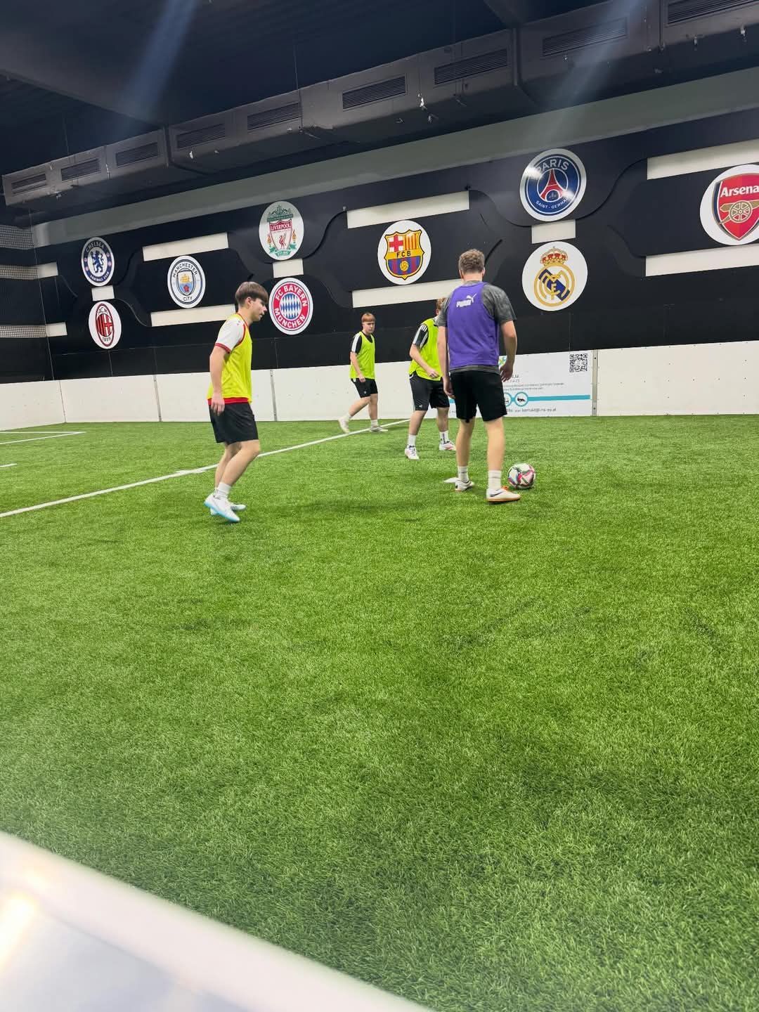 Vier Fußballspieler üben auf einem Rasenplatz in einer Halle, einer Spieler bereitet sich darauf vor, den Ball zu schießen. Die Spielfläche hat Logos an den Wänden.