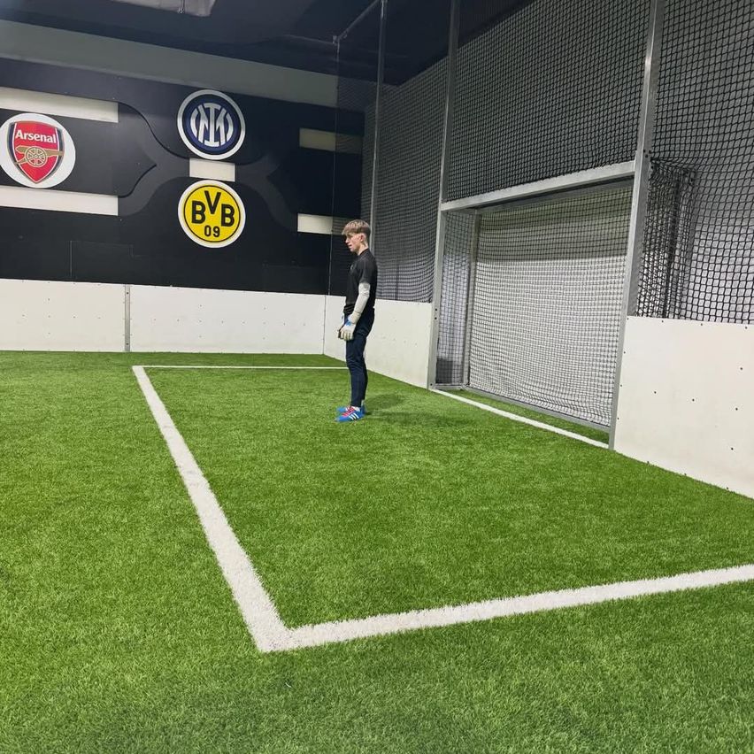 Ein junger Junge steht auf einem grünen Feld, trägt ein schwarzes Shirt, blaue Sneaker und Handschuhe. Hinter ihm befindet sich ein Fußballtor und eine Wand mit Logos für Arsenal, Inter Mailand und Borussia Dortmund.