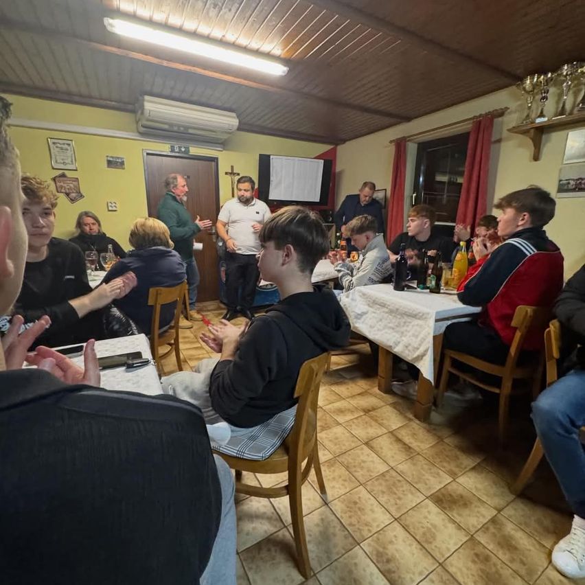 Eine Gruppe von Menschen versammelt sich in einem Raum, einige sitzen um Tische herum, andere stehen. Ein Mann scheint mit einem anderen Mann zu sprechen. Der Raum hat geflieste Böden und Wände, die mit verschiedenen Gegenständen geschmückt sind.