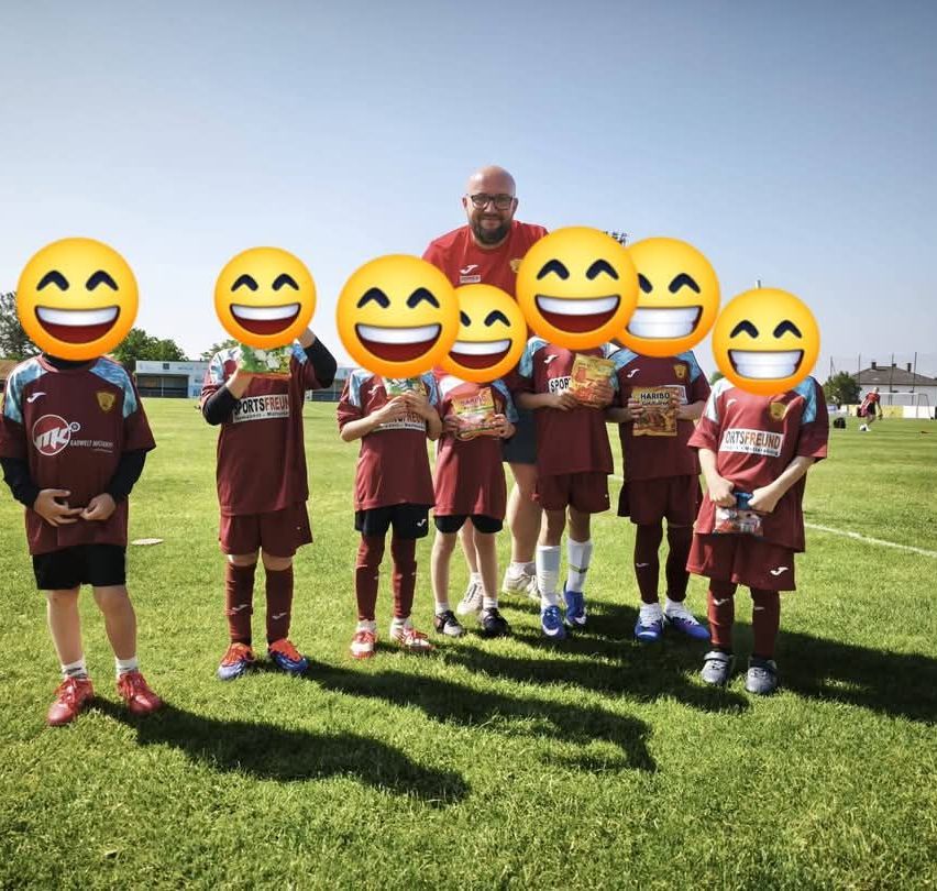 Eine Gruppe junger Fußballer mit lächelnden Gesichtern posiert für ein Foto. Der Mann in der Mitte hält Snacks. Die Kinder tragen dunkelrote Trikots.