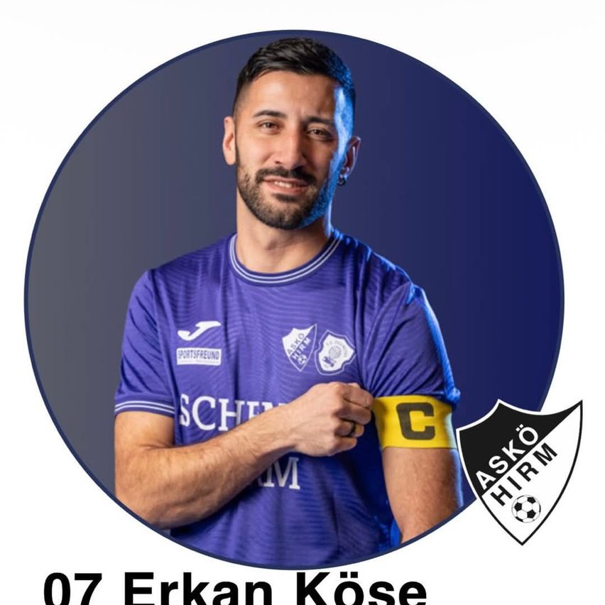 Ein lächelnder Mann in einem violetten Fußballtrikot mit der Nummer 07, Erkan Kose, zeigt auf seinen Arm, der ein gelbes Armband mit dem Buchstaben C trägt.