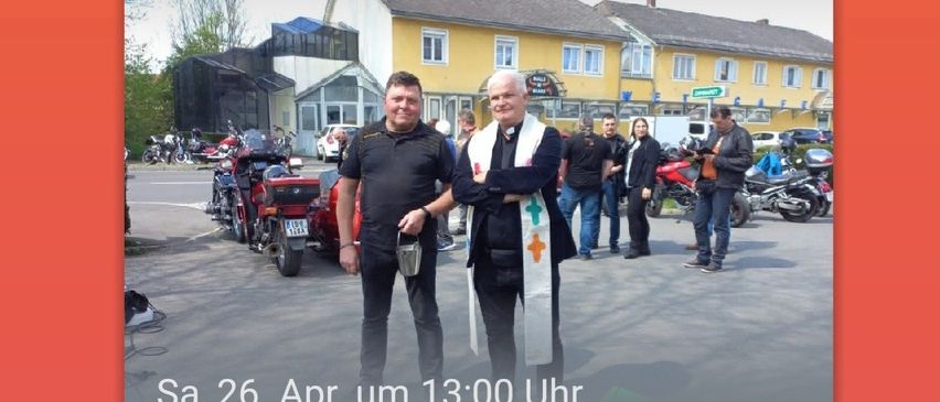 Zwei Männer stehen vor einem gelben Gebäude mit geparkten Motorrädern im Hintergrund. Der Mann auf der linken Seite hält einen Becher. Der Mann auf der rechten Seite hält ein weißes Tuch mit einem Kreuz. Das Bild hat einen Wasserzeichen mit dem Text 'Sa, 26. Apr. um 13:00 Uhr Motorradsegnung!'.