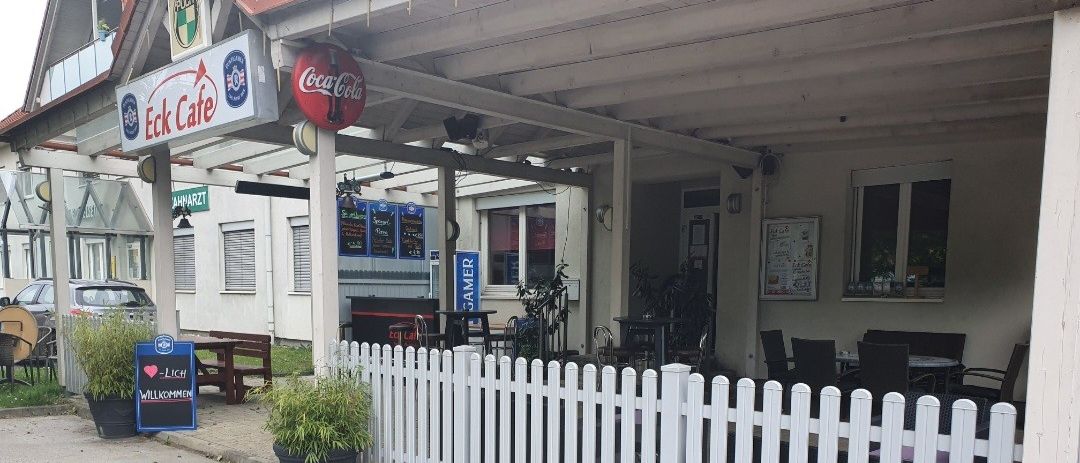 Außenbereich des Eck Cafe mit Coca-Cola-Schild, weißer Picket-Zaun, Tischen und Stühlen, Topfpflanzen und einem Willkommensschild.