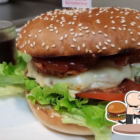 Ein Burger mit Sesamkörnern auf dem Brötchen, belegt mit geschmolzenem Käse, Tomaten, Salat und Speck. Ein kleines Schild mit dem Namen 'Bad Radkersburg' ist auf dem Burger.