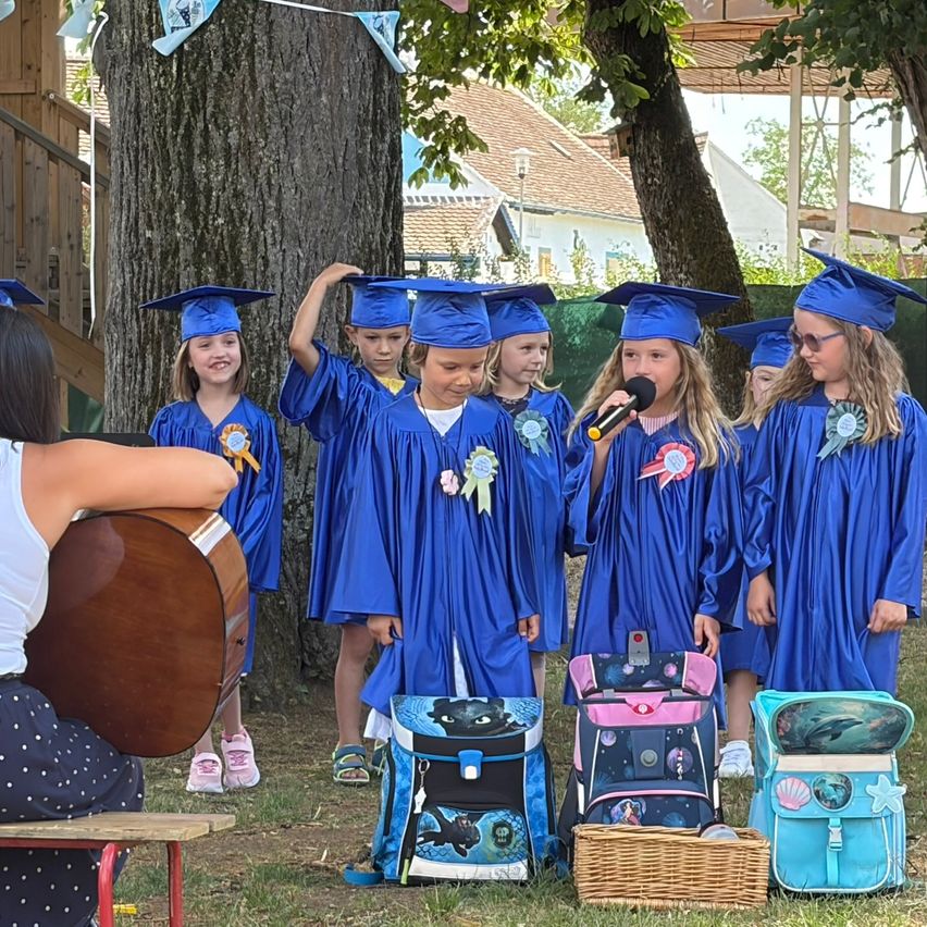 A group of children in blue graduation gowns and hats sing with a microphone while a woman holds a guitar. They have colorful backpacks and are surrounded by trees and houses.